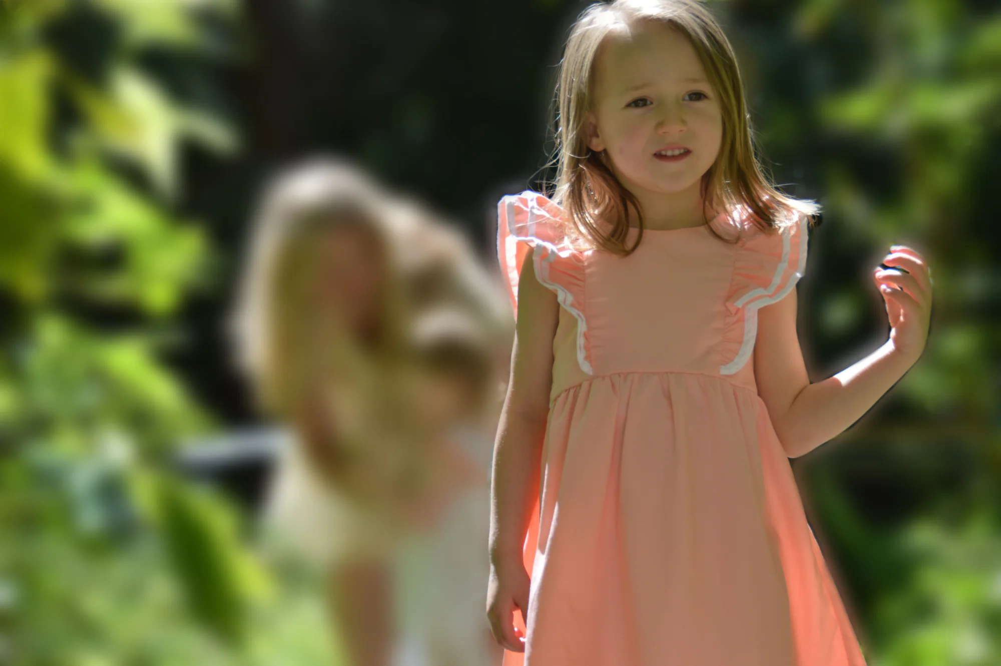 Young girl stands before softly blurred adult and leafy garden.