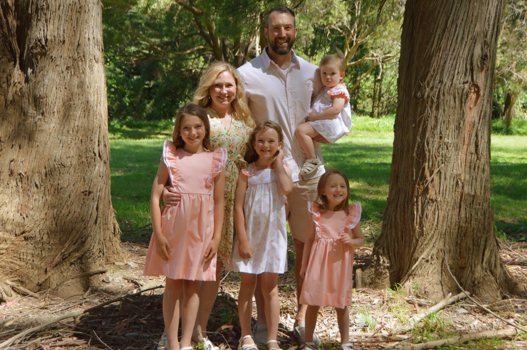 Parents and four daughters pose between two trees in sunny woodland.