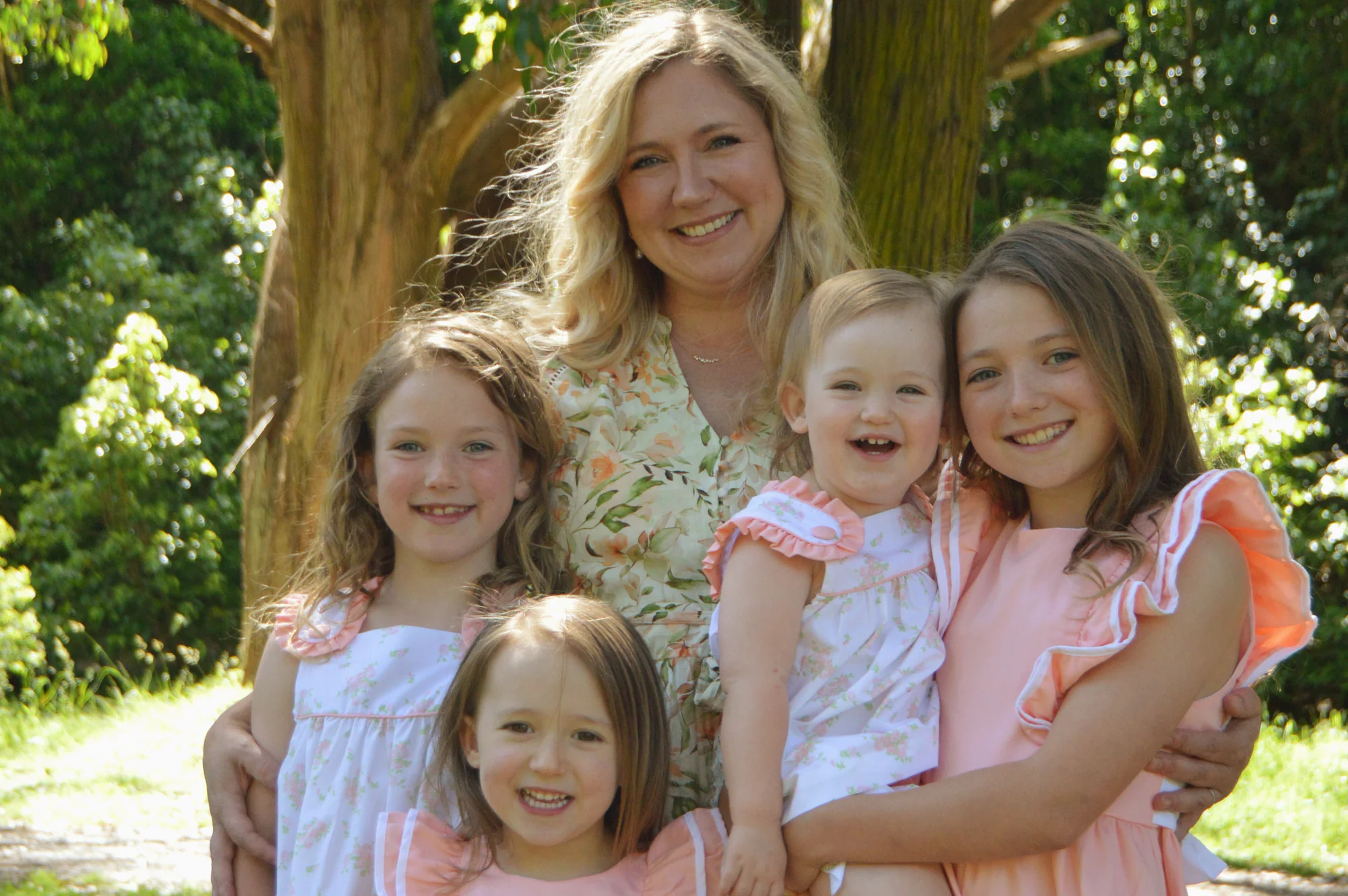 Mother and four daughters smile together beneath sunlit trees.