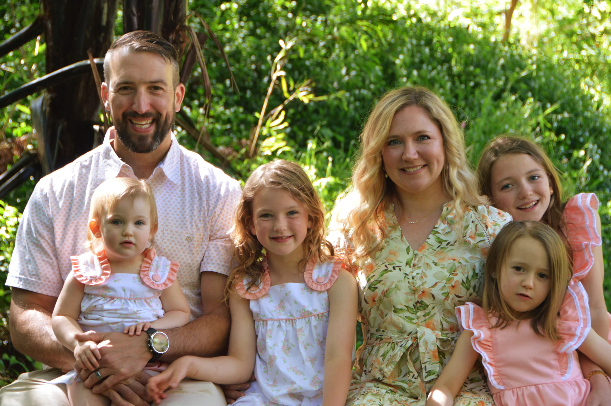 Parents sit closely with four daughters beneath leafy trees.