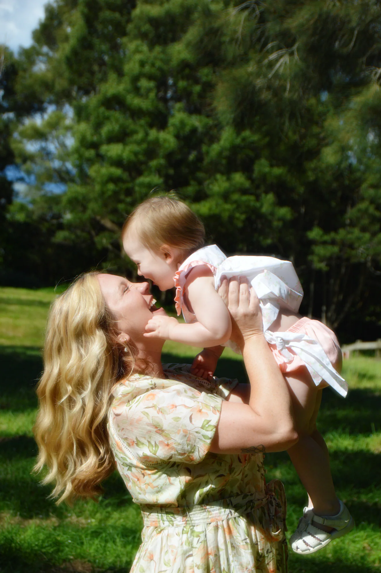 Mother lifts laughing toddler close beneath a sunny blue sky.