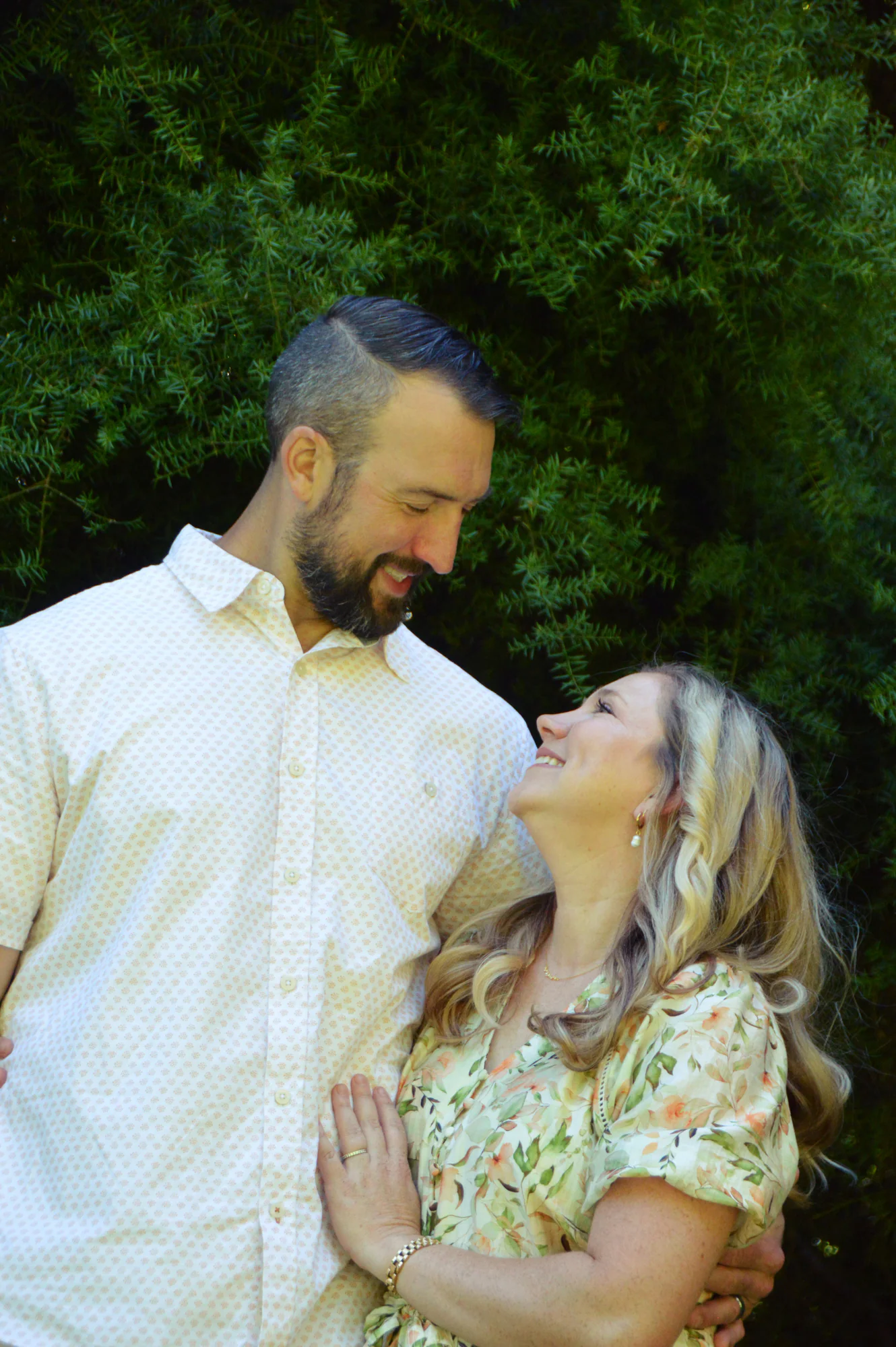 Couple smile at each other against dense green foliage.