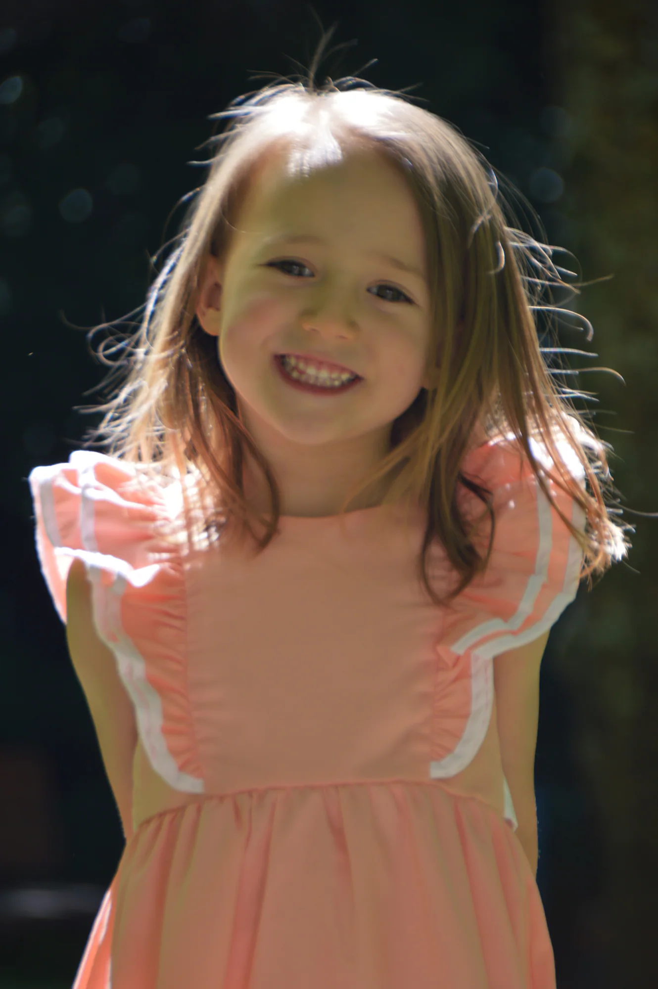 Smiling young girl in pink dress stands against dark foliage.