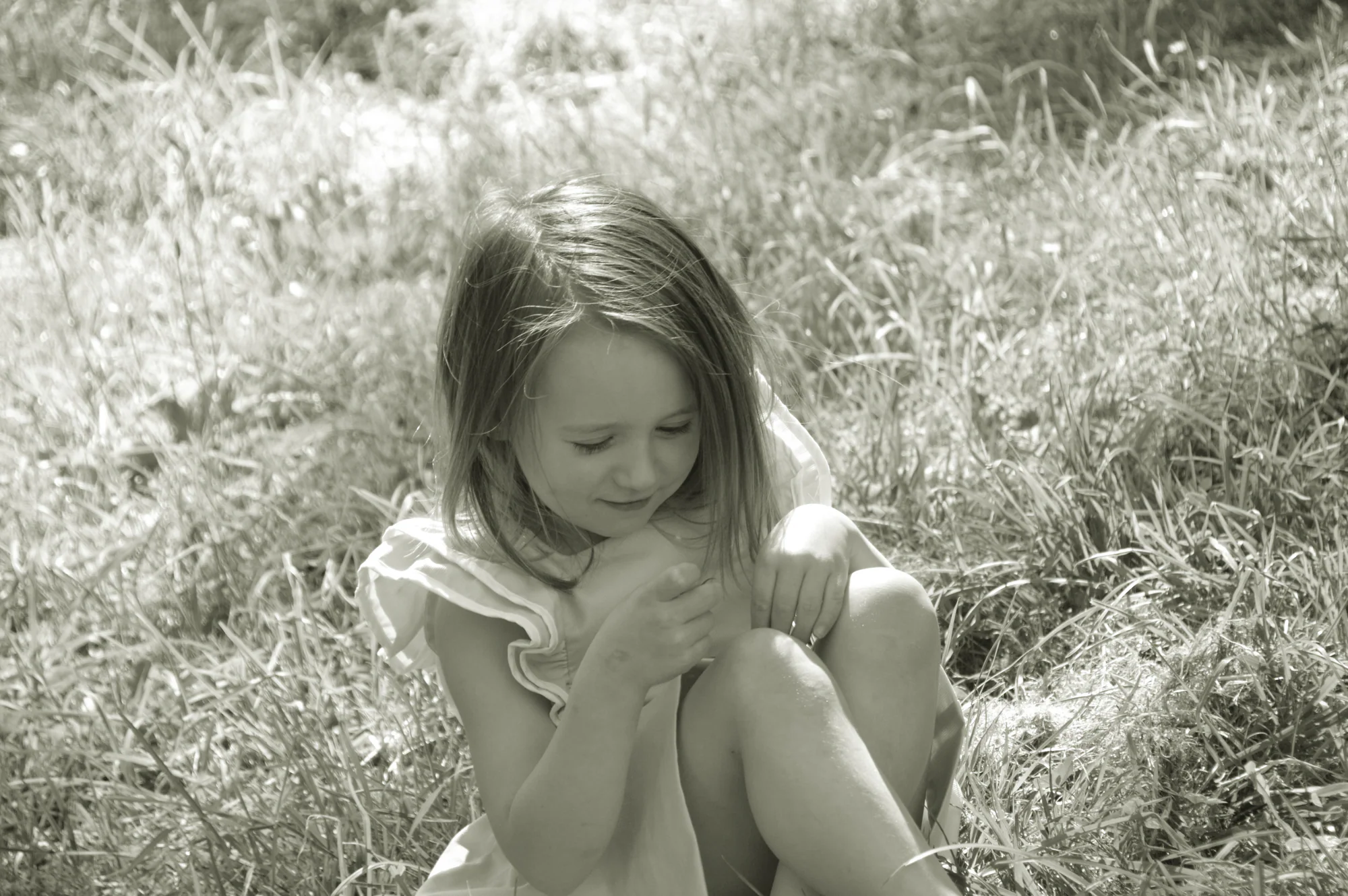 Seated girl looks down thoughtfully amid sunlit grass in monochrome.