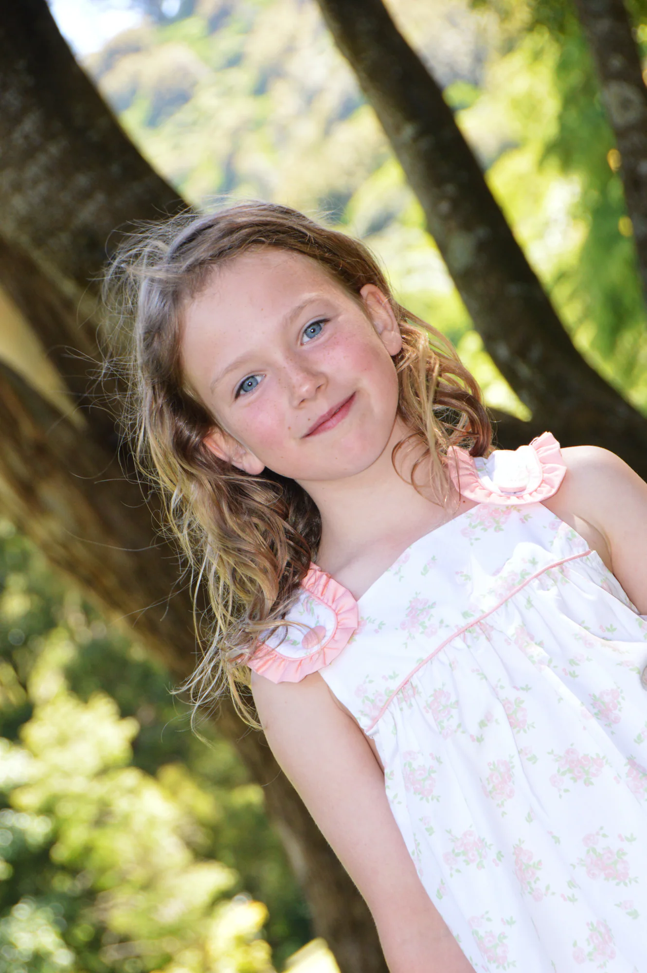Smiling girl in floral dress tilts playfully against leafy woodland.