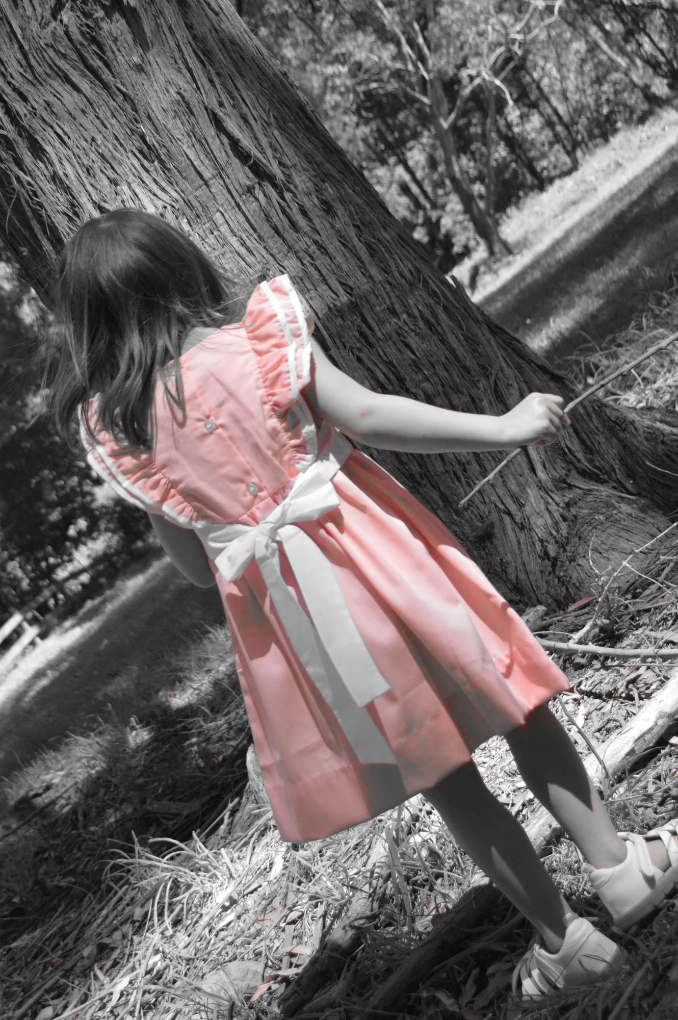 Young girl in pink dress reaches toward tree in selective color.