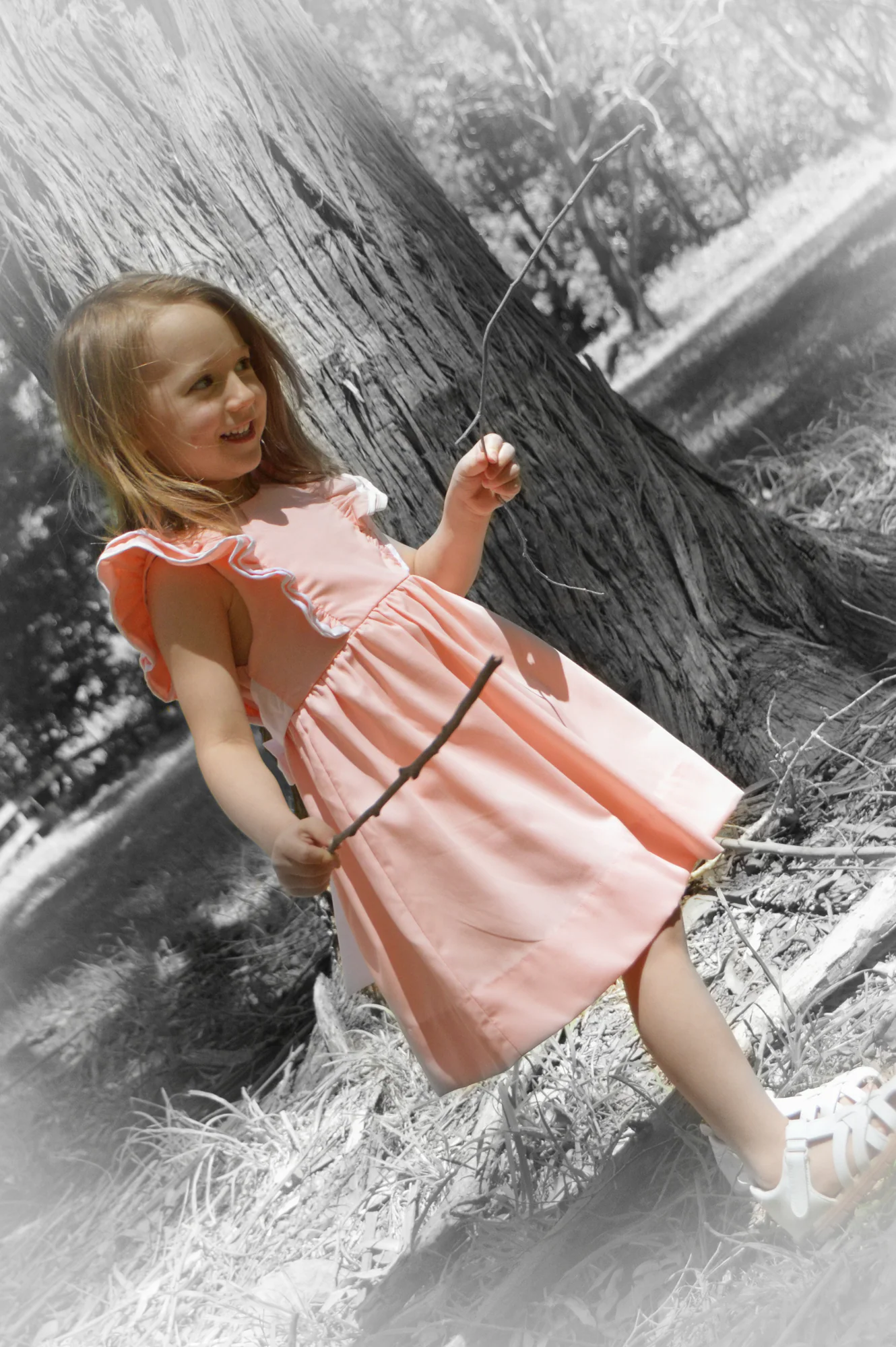 Smiling girl in pink dress leans beside tree in selective color.